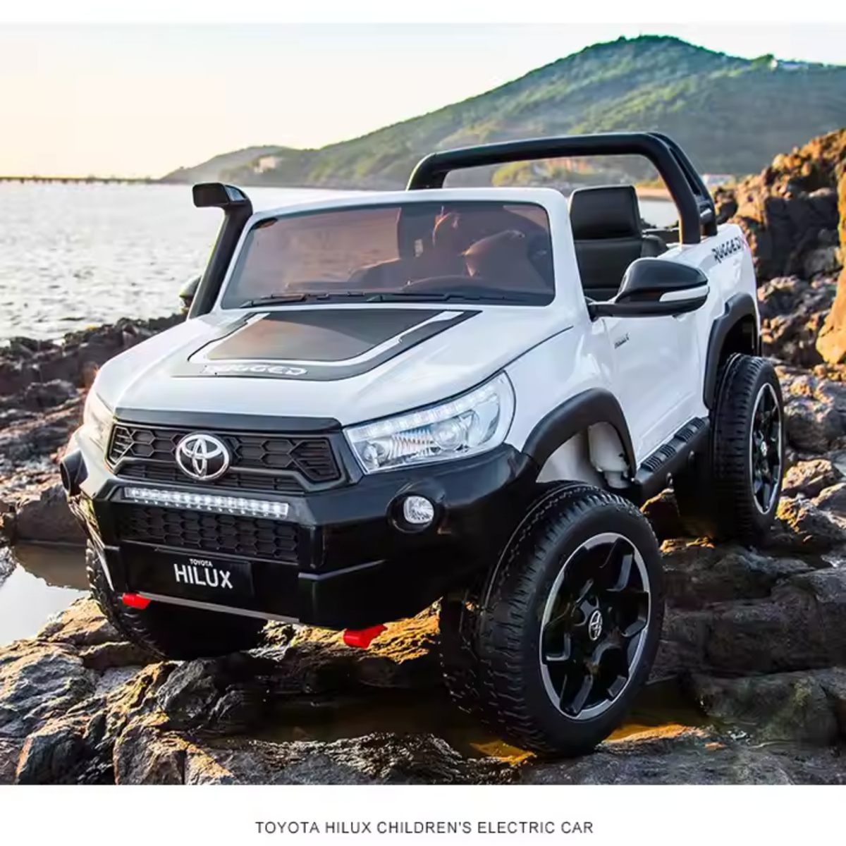 White licensed Toyota Hilux 24V kids ride-on car with 4x4, LED lights, and rugged off-road tires on rocky terrain