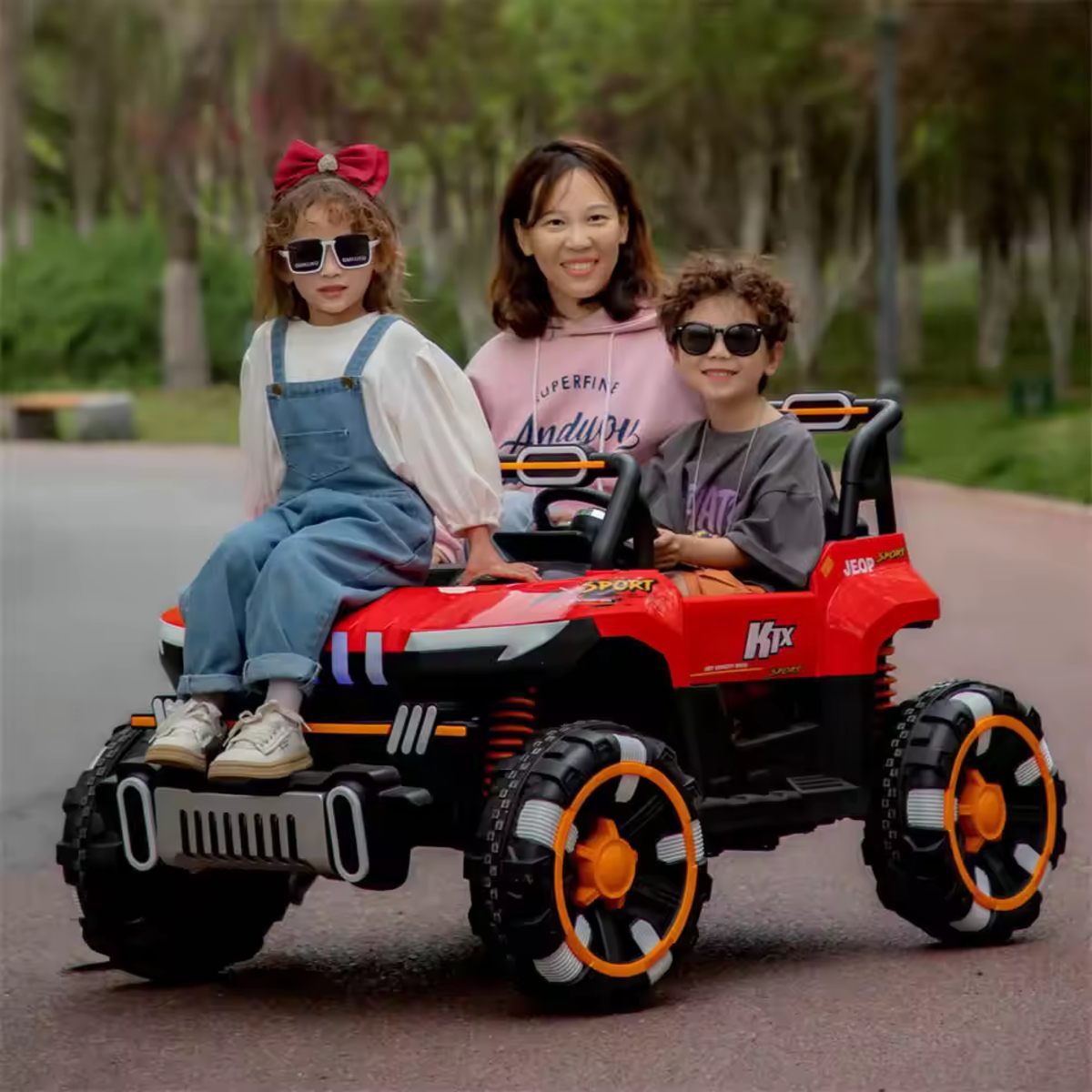 Two kids and an adult enjoying a red 24V kids UTV ride-on buggy with black and orange wheels outdoors