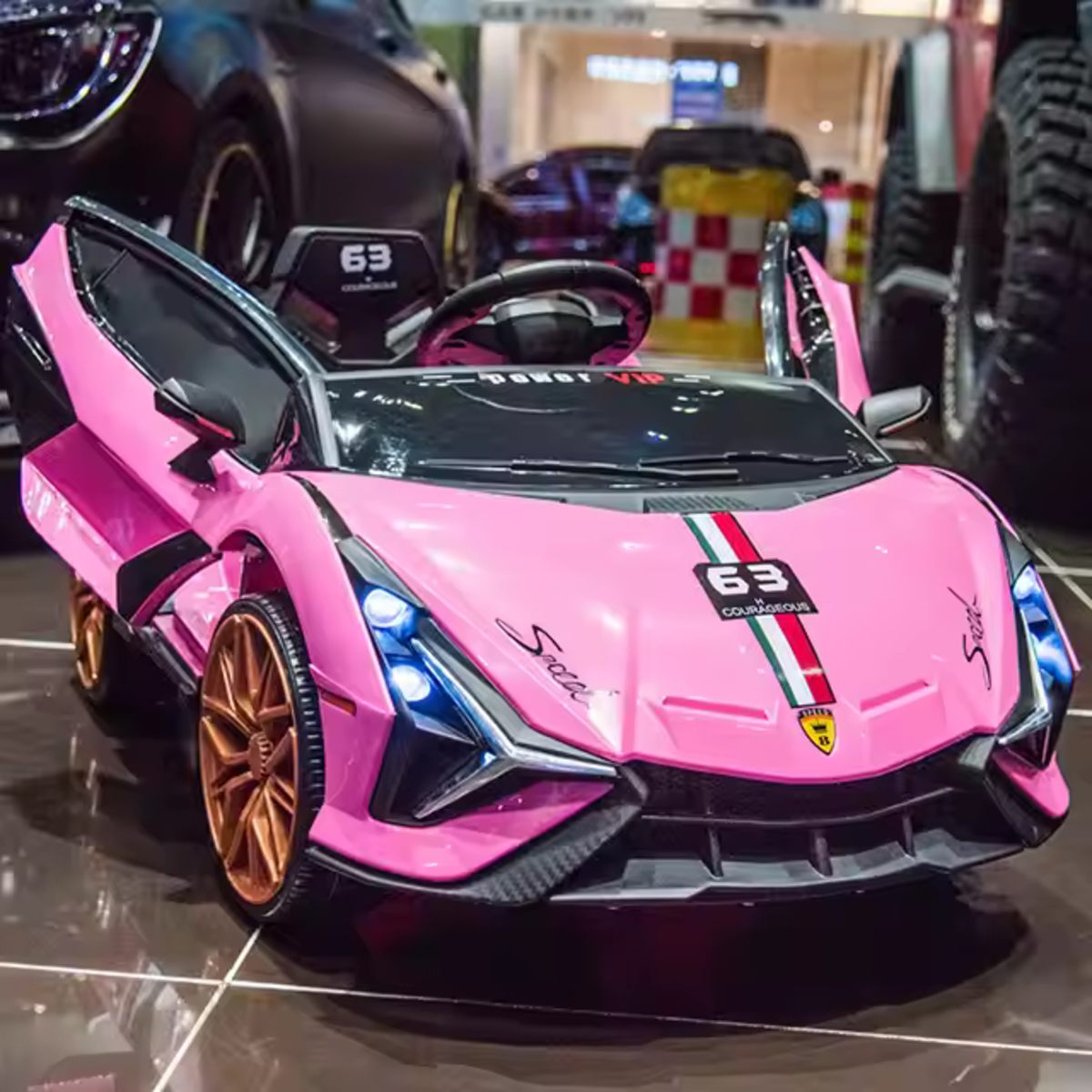The MC Wholesalers 12v Lamborghini Inspired ride on race car in pink features butterfly doors, gold wheels, and racing decals. Displayed indoors on a shiny floor, it's shown with other vehicles in the background.